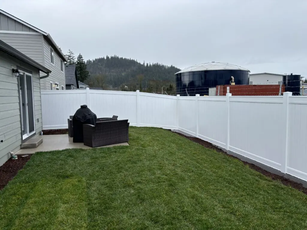 backyard with freshly installed white vinyl privacy fence surrounding green lawn and outdoor patio furniture with covered grill next to modern house under overcast sky, showcasing Timberhill Fence & Deck residential fencing and outdoor living solutions