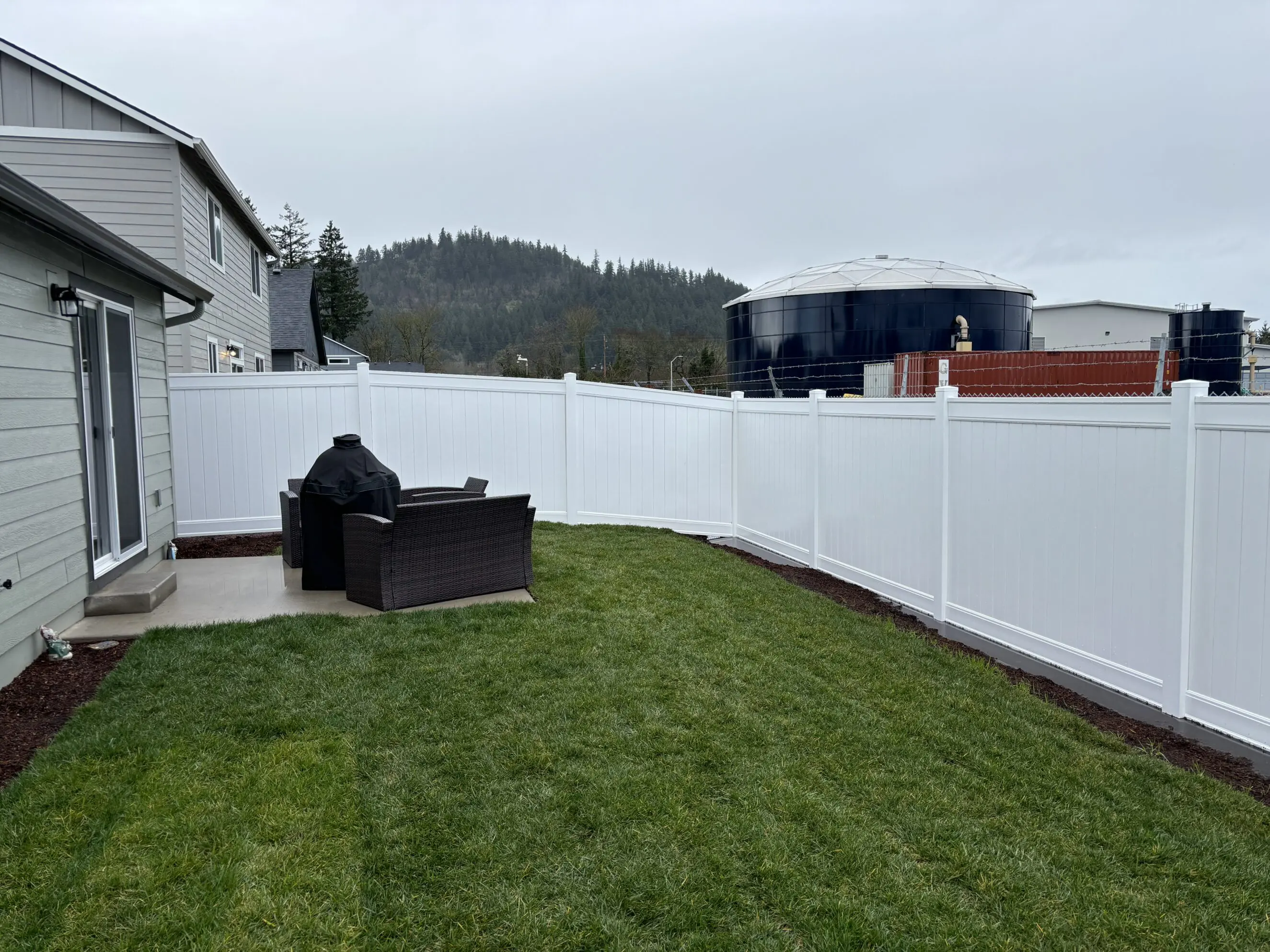 IMG_6016 Backyard with freshly installed white vinyl privacy fence surrounding a green lawn patio area with outdoor seating and a covered grill, showcasing Timberhill Fence & Deck's quality fence installation services.
