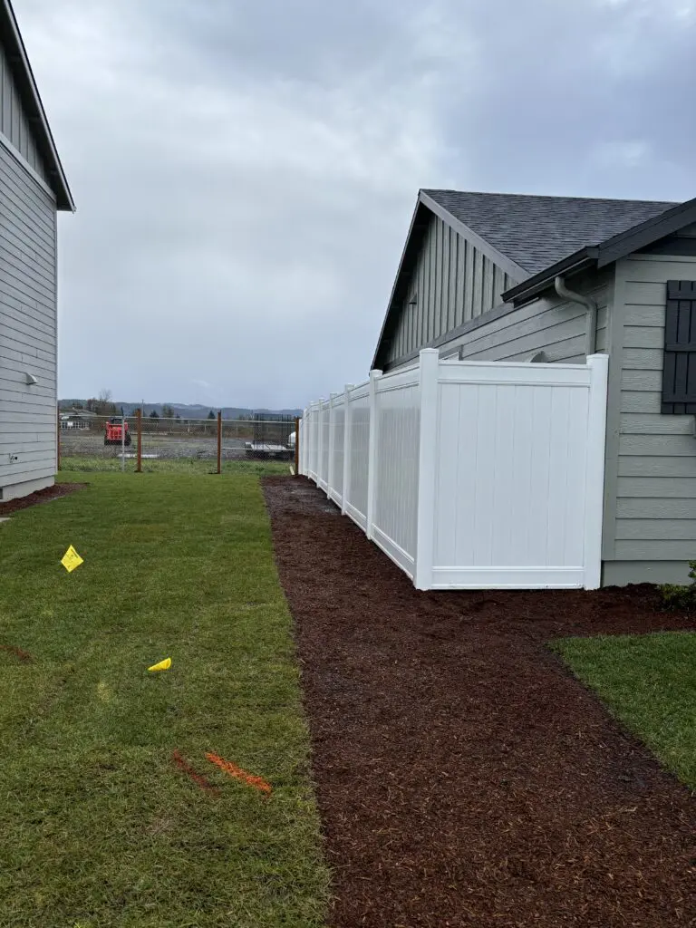 Residential backyard with newly installed white vinyl privacy fence, fresh mulch bed along the fence line, and green lawn between two houses under overcast sky, showcasing Timberhill Fence & Deck professional fencing and landscaping services.