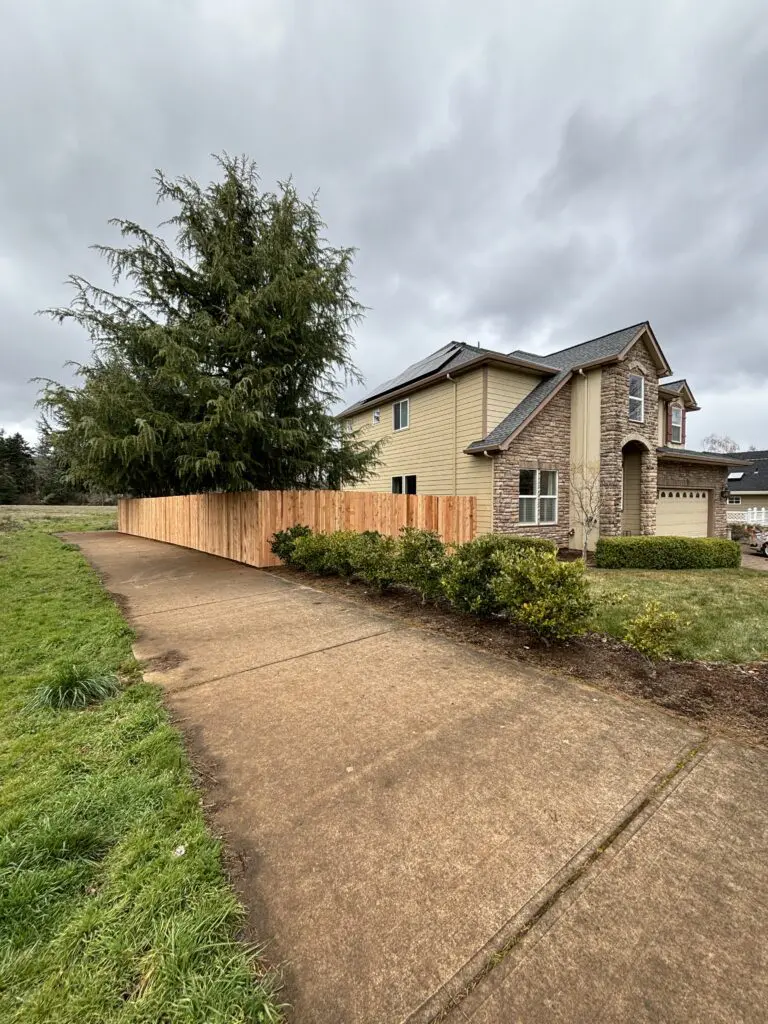 Two-story suburban home with stone and beige siding, solar panels on roof, surrounded by fresh wooden privacy fence, landscaped shrubs, large evergreen tree, and concrete sidewalk under cloudy sky.
