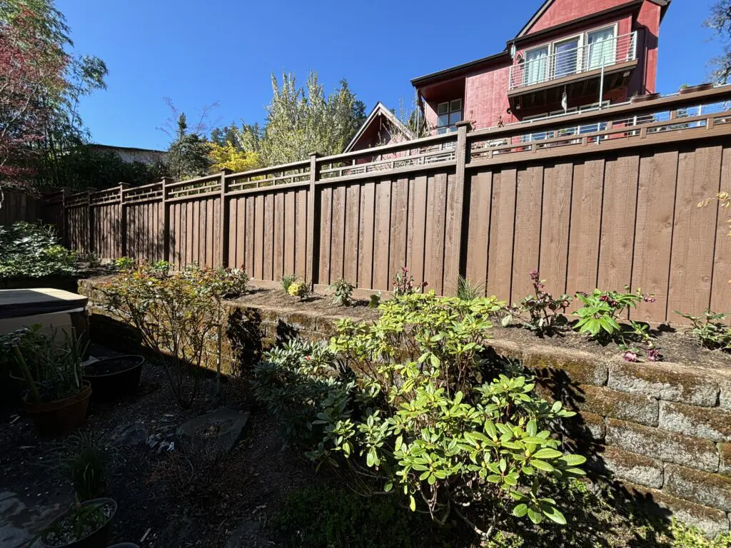 Backyard garden with a newly installed brown wooden privacy fence and stone retaining wall, surrounded by shrubs and plants under clear blue sky, showcasing Timberhill Fence & Deck's custom fence installation services.