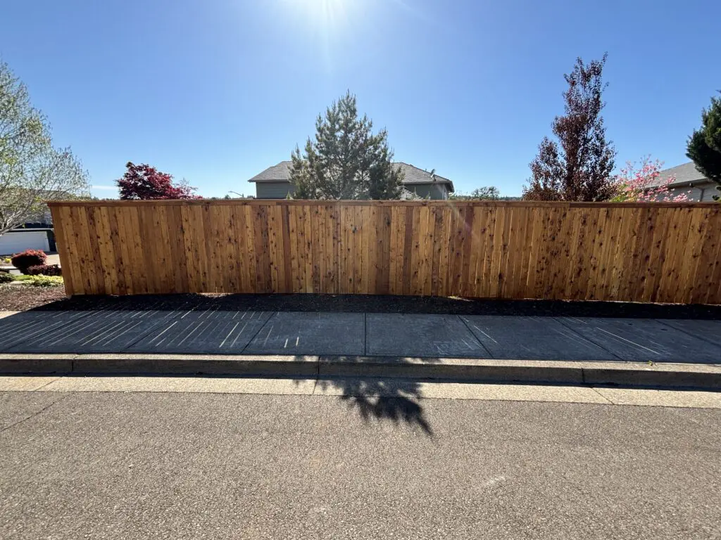 Newly installed natural cedar wood privacy fence along a residential sidewalk under a clear blue sky, showcasing quality craftsmanship by Timberhill Fence & Deck.