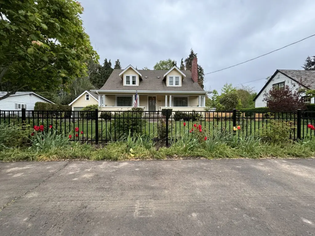 Front view of a beige Timberhill Fence & Deck home with a black metal fence, green lawn, red and yellow tulips, and an American flag under a cloudy sky.