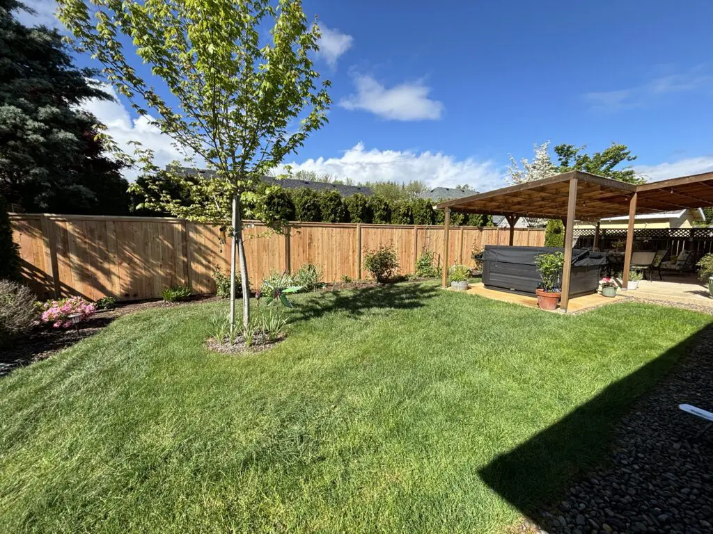 backyard with freshly installed cedar fence, green lawn, young tree, landscaped flower beds, and covered patio with hot tub under clear blue sky at Timberhill Fence & Deck residential property