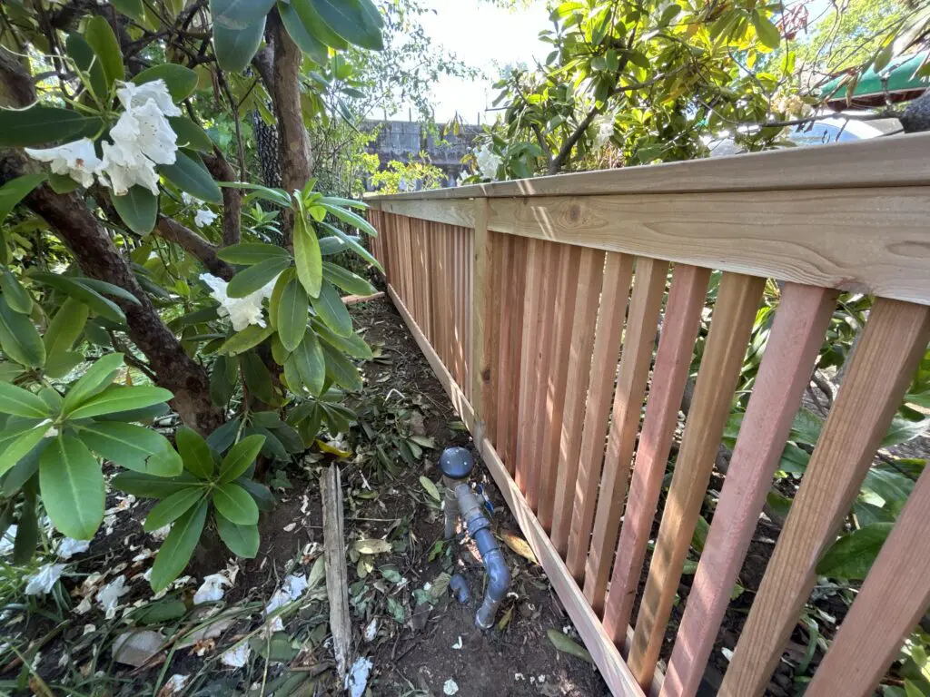 Wooden fence installed along garden bed with green plants and white flowers, highlighting Timberhill Fence & Deck craftsmanship and outdoor landscaping.