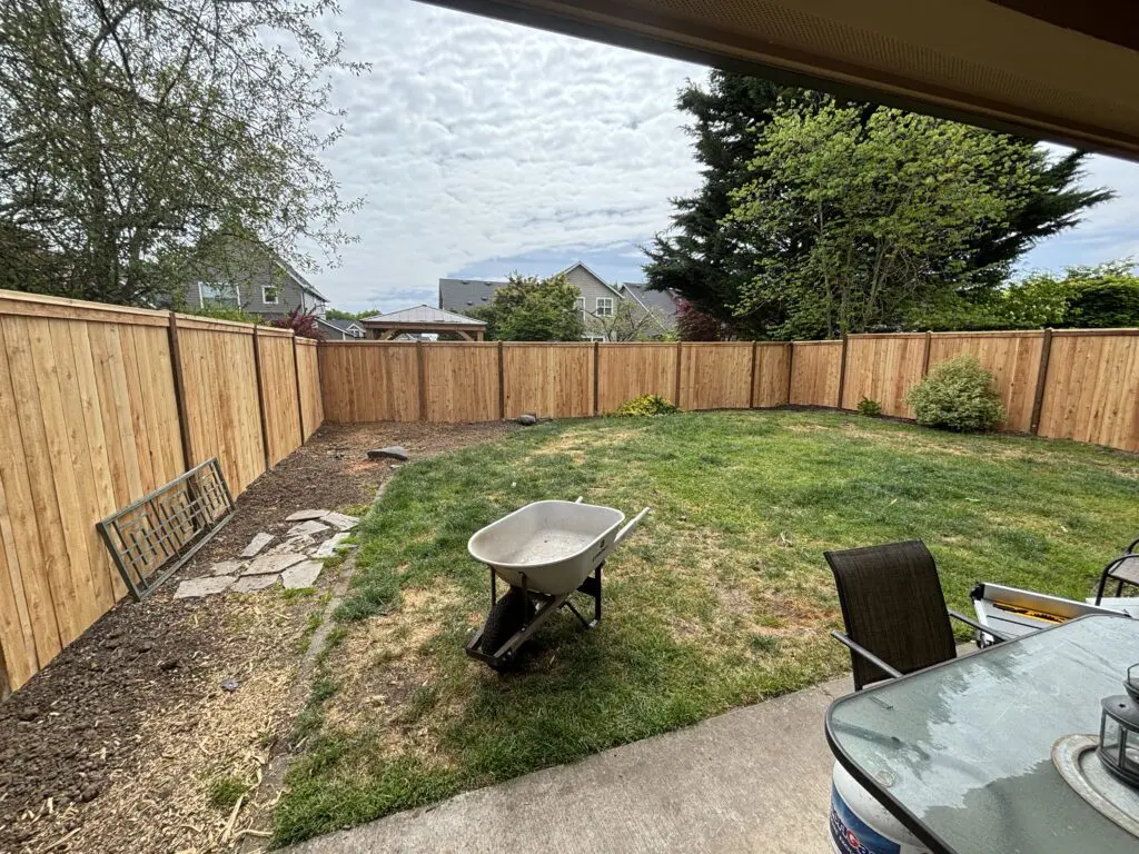 backyard with freshly installed wooden fence, patchy grass lawn, wheelbarrow on grass, patio area with outdoor furniture, trees and neighboring houses in the background