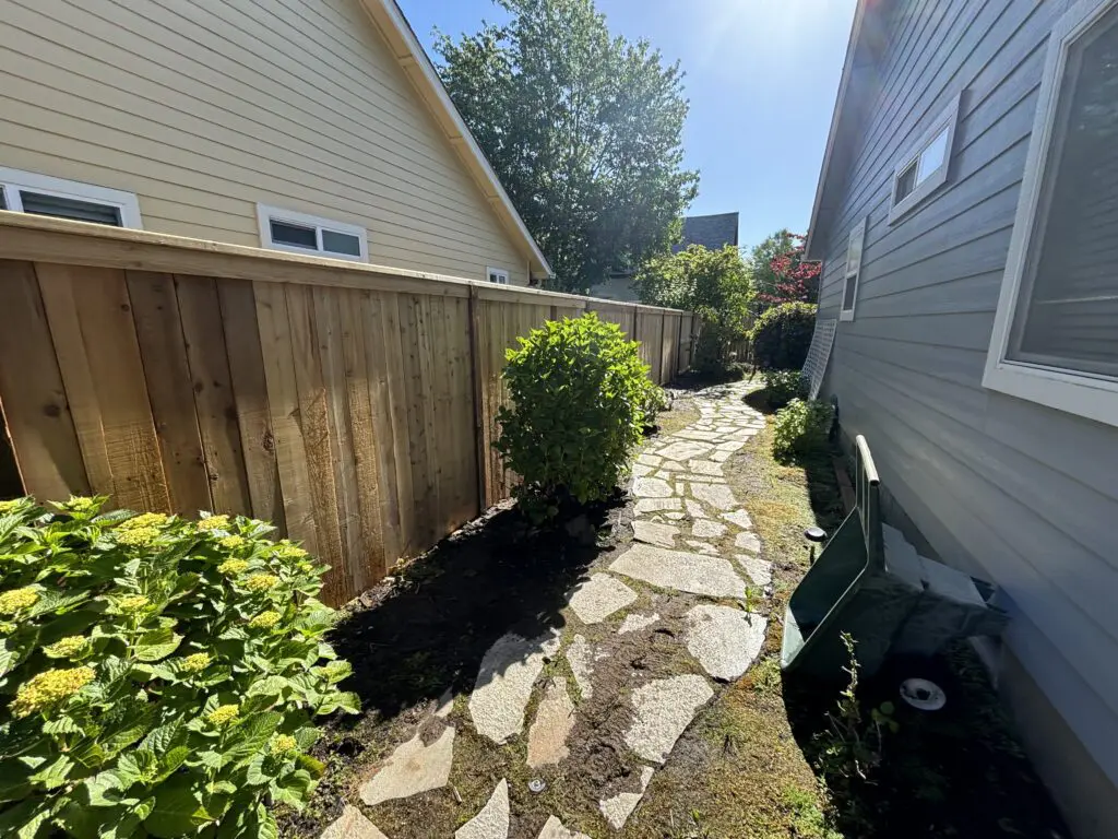 Stone pathway bordered by green shrubs and a wooden fence along the side of a residential house, showcasing Timberhill Fence & Deck's craftsmanship in outdoor landscaping and deck design.