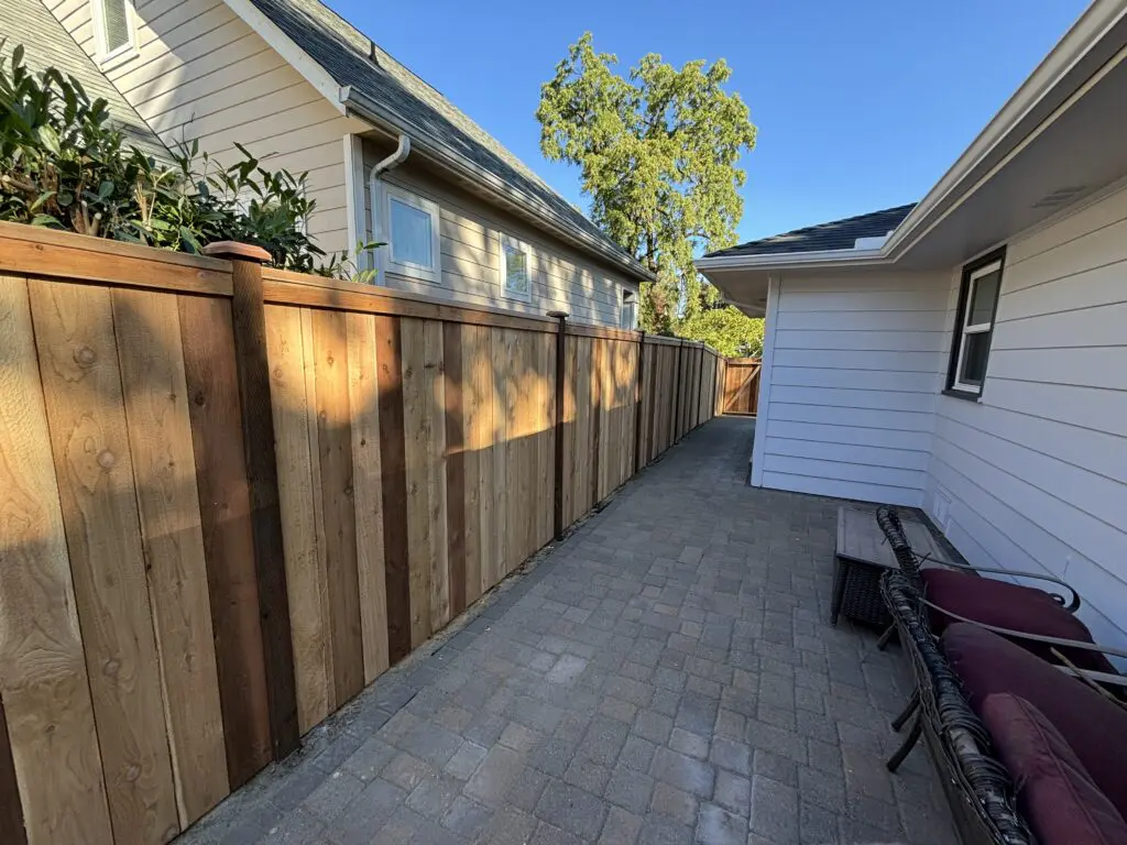 narrow backyard patio with new wooden fence, paved stone walkway, and white siding house exterior under clear blue sky, showcasing Timberhill Fence & Deck quality craftsmanship and outdoor living space enhancement