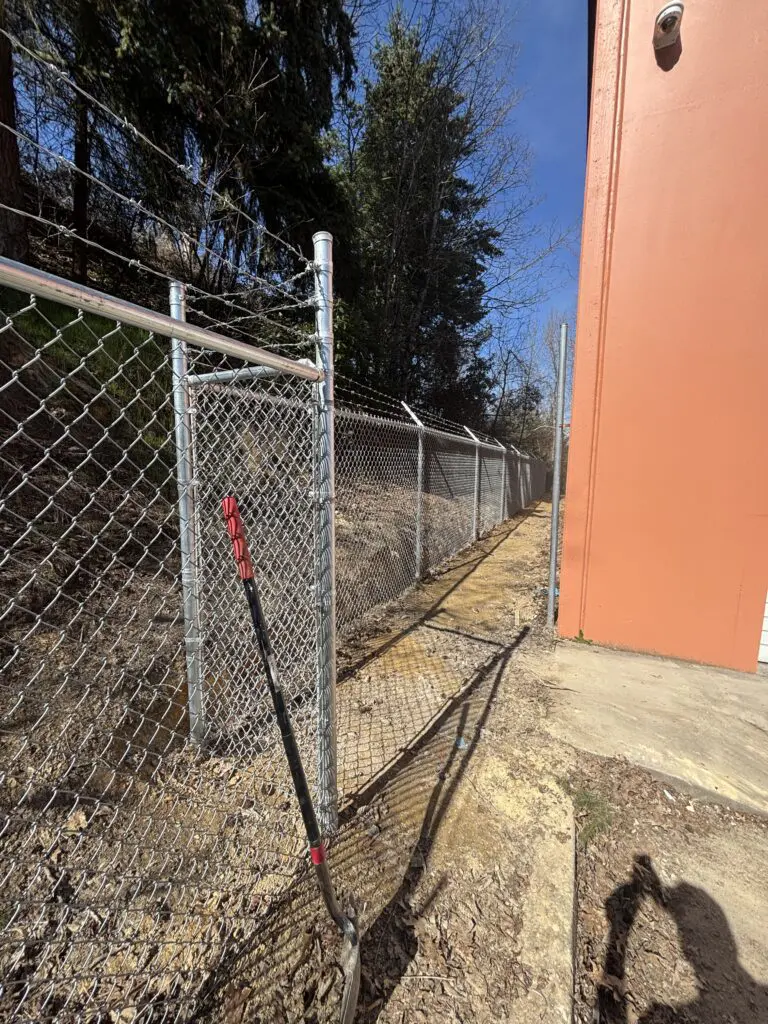 Chain link fence installation along a property line next to an orange building wall with a digging tool leaning against the fence and wooded area in the background under a clear blue sky, illustrating Timberhill Fence & Deck's professional fence construction services.